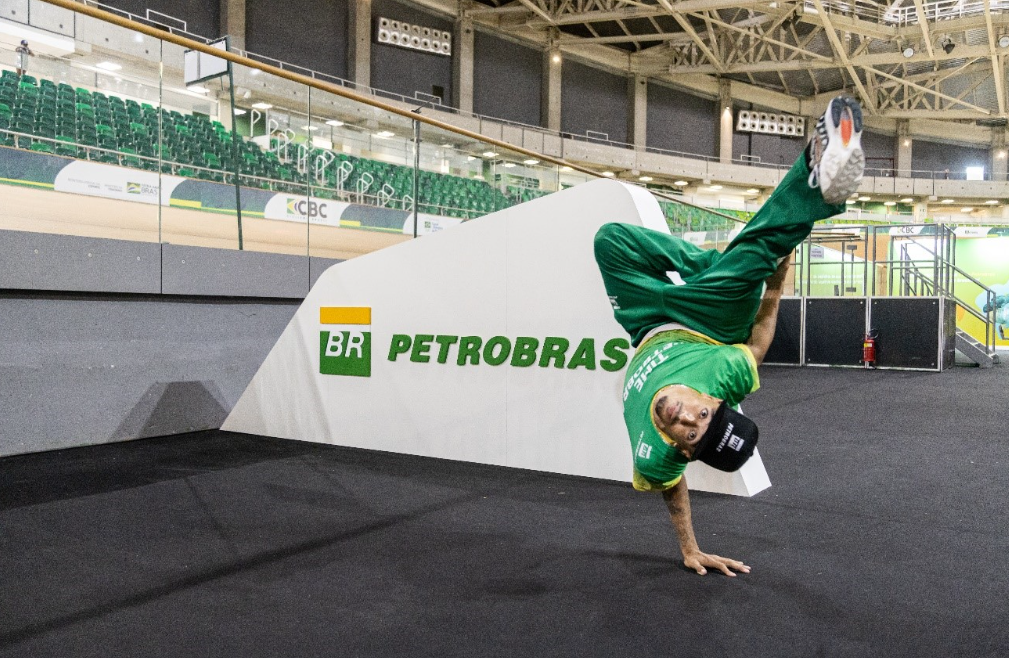 Luan San, atleta de Breaking, do Time Petrobras fazendo uma manobra, com a mão apoiada no chão e as pernas no ar, num ginásio, com a marca Petrobras ao fundo